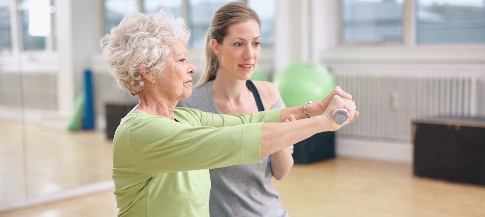 An older woman in a green shirt performs an exercise with hand weights, assisted by a younger woman in a skilled nursing fitness studio.