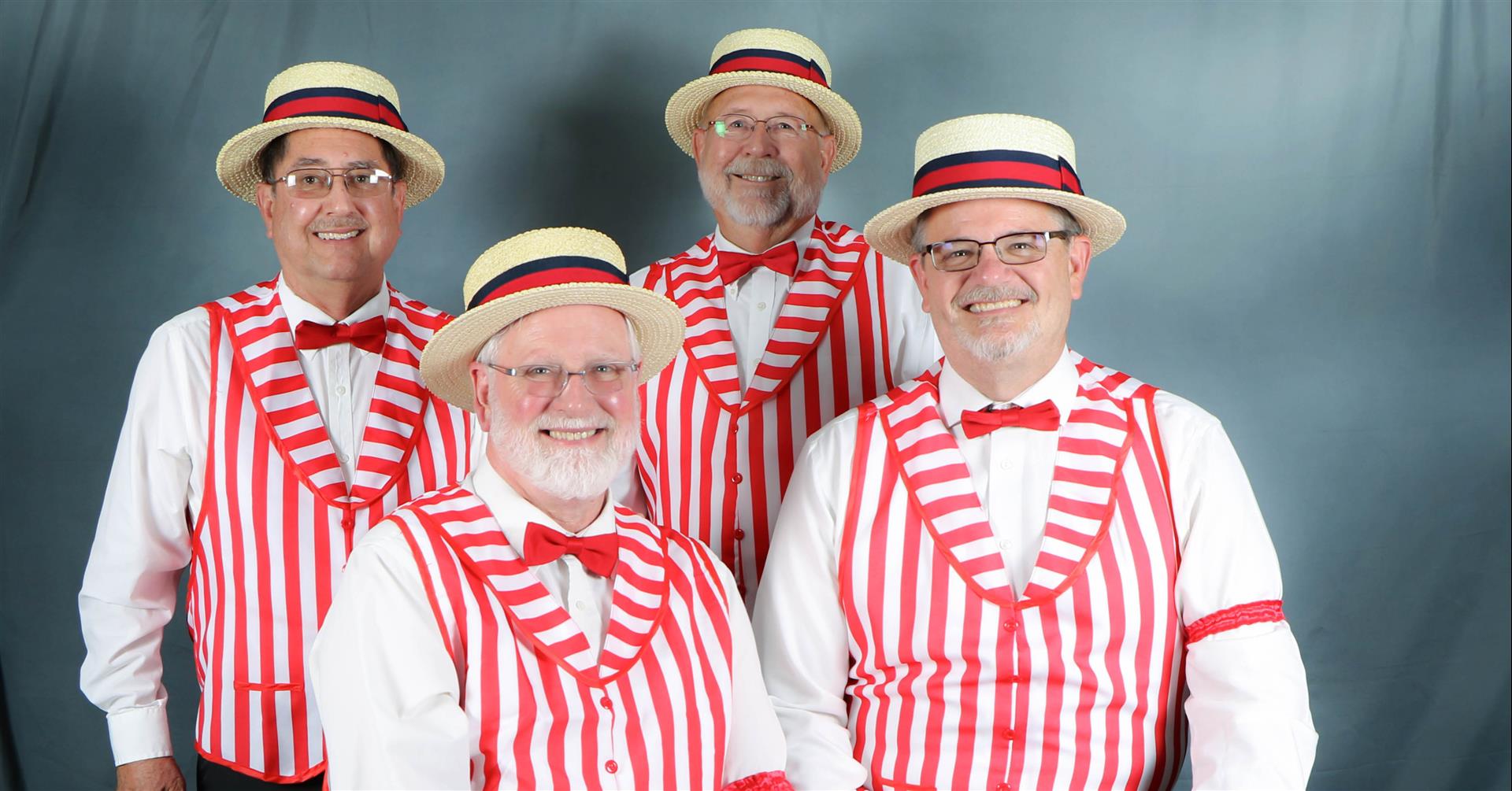 Four men dressed in white shirts, red bow ties, red and white striped vests, and straw hats with red and blue bands stand and pose against a neutral background, forming a charming barbershop quartet.