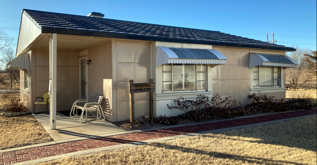 Single-story Lustron Home with light-colored siding and a dark roof. The house features a covered entryway with two chairs, awnings over the windows, and a small sign near the front door. Dry grass surrounds this charming property in Great Bend, Kansas.