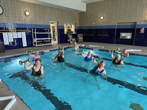 A group of eight people participates in a water aerobics class, lifting foam dumbbells while standing in a swimming pool. The pool area, located at Wichita Presbyterian Manor, has a tiled wall and several safety railings in view, exemplifying the active independent senior living lifestyle.