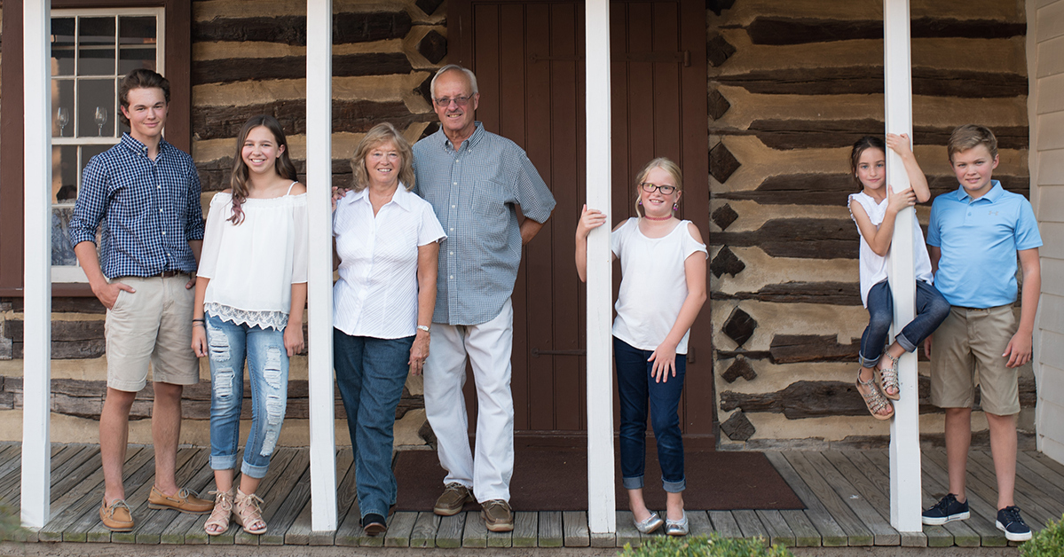 A group of six people, including Nana, PopPop, two adults and four children, standing and sitting on the porch of a log cabin-style building.