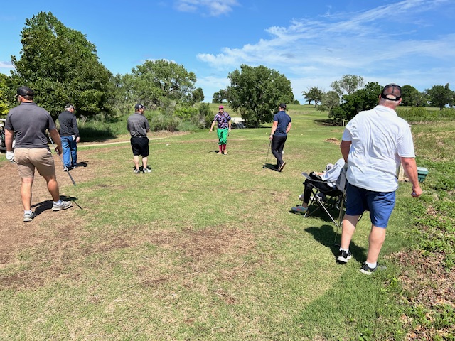 Several people on the golf course at Presbyterian Manor in Arkansas City stand in various poses holding golf clubs, with trees and a clear blue sky in the background. They're participating in the Golfin' for Good Sam event.