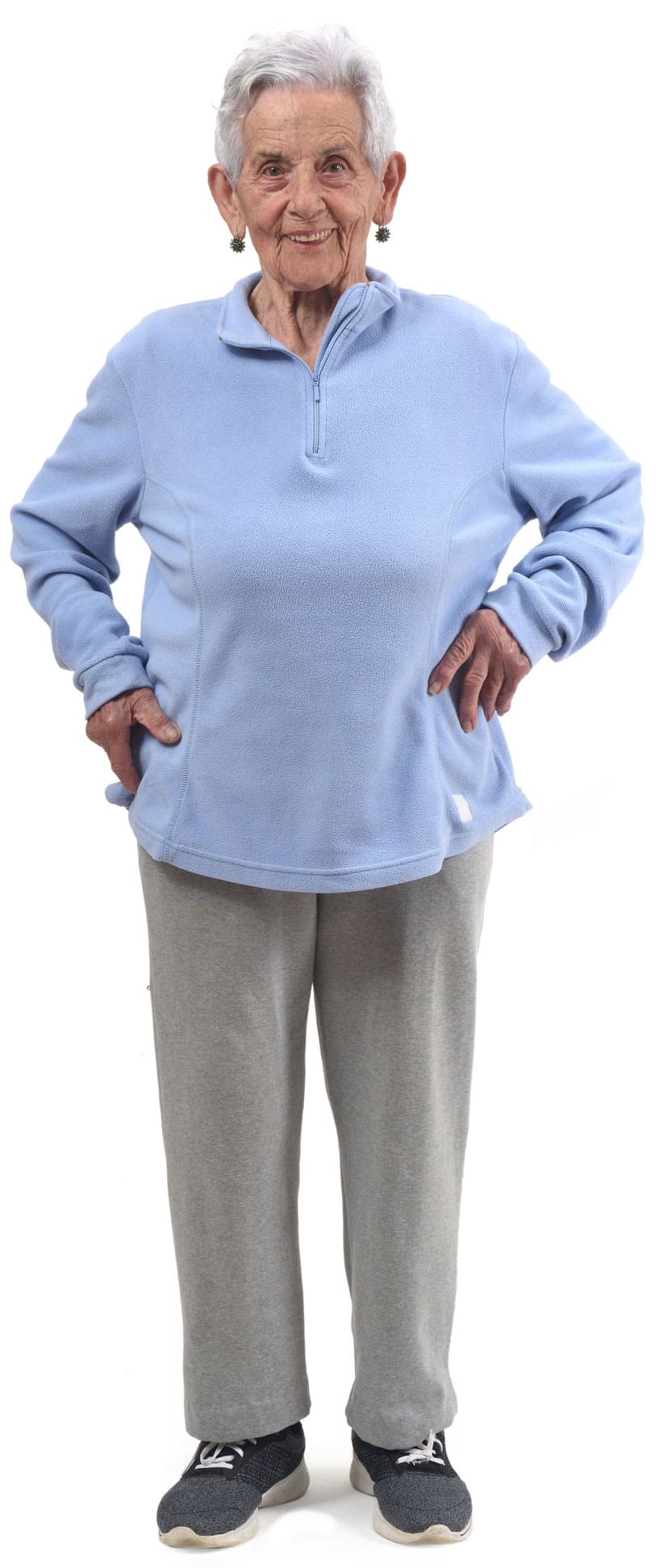 Elderly person with gray hair, wearing a blue pullover, gray pants, and black sneakers, standing confidently with hands on hips against a white background, ready for short-term rehab.