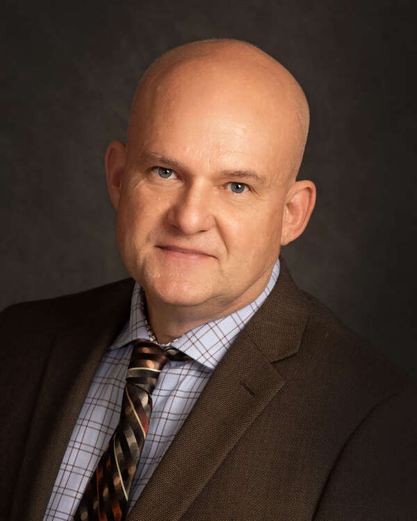 A bald man in a brown suit and patterned tie, wearing a checkered shirt, poses against a dark background.