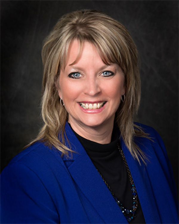 A woman with blond hair, wearing a blue blazer over a black top, smiles at the camera against a dark background.