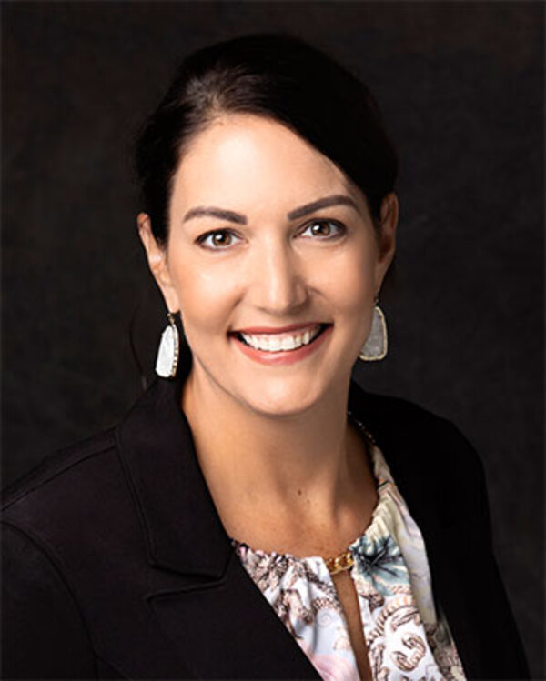 A woman with dark hair and wearing a black blazer and patterned blouse smiles at the camera against a dark background. She also wears drop earrings.