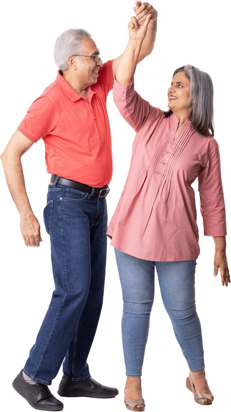 An older man in a red polo shirt and jeans is holding hands and dancing with an older woman in a pink top and jeans. They are smiling and looking at each other, enjoying their time together at senior living in Colorado.